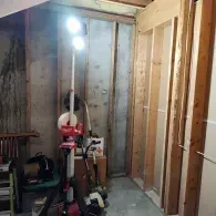 A bright, portable Milwaukee LED work light stands in a room undergoing construction with exposed wooden wall framing.