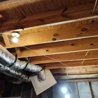 Exposed ceiling joists in an unfinished basement, featuring a mounted light fixture and flexible silver HVAC ducts.