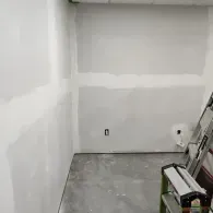 An indoor corner featuring bare, unfinished drywall with visible joint compound seams and a folding ladder on the floor.