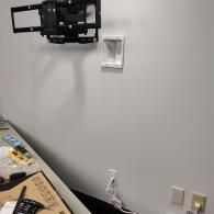 A TV wall mount bracket installed on a white wall above an in-wall cable management box, with electrical outlets below.