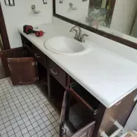 A bathroom vanity with a white countertop and sink, featuring missing cabinet doors and a power drill on the counter.