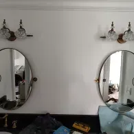 Two oval wall mirrors with ornate light fixtures above each, mounted on a white wall above a dark vanity.