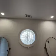 A bathroom wall with two oval mirrors flanking a central round window, under two recessed ceiling lights and a vent.