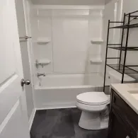 A bathroom featuring a white bathtub and shower unit, a white toilet, and a dark metal shelving unit on the right.