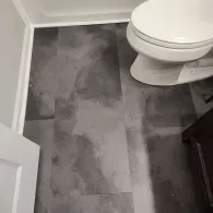 A white toilet sits on a floor covered in dark gray, mottled, rectangular luxury vinyl plank flooring in a bathroom.