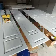 Light gray painted doors laid flat on workbenches in a garage workshop with a paint can resting on one door.