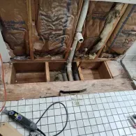 An open wall section and floor cutout revealing plumbing pipes, insulation, and wooden joists in a tiled bathroom.