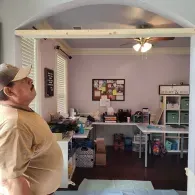 A person in a beige shirt looks up at a wooden beam newly installed across an arched doorway leading to a home office.