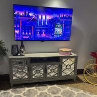 A wall-mounted TV showing a colorful city scene above a gray, four-door mirrored console table on a patterned rug.