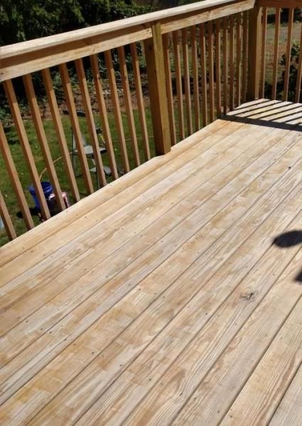 A wooden deck with a railing, featuring light brown, weathered planks viewed from above looking out toward a grassy yard.