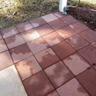 A patio made of square, red concrete pavers partially wet from rain, situated next to grass and a white porch post.