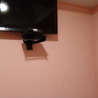 A black flat-screen television mounted on a plain, light pink wall with a small black shelf holding electronic equipment.