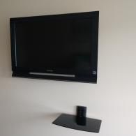A black flat-screen television mounted on a plain white wall above a small, black floating glass shelf.