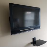A black television mounted on a beige wall above a small, black floating shelf.