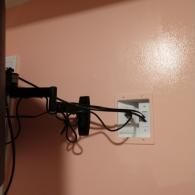 A black TV wall mount extends from a light pink wall, with cables running into a recessed white wall box.