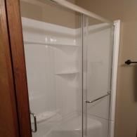 A white walk-in shower stall with built-in shelves and a bench, enclosed by clear glass sliding doors.
