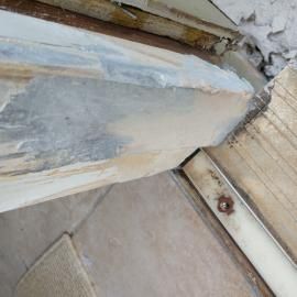 Close-up of a damaged, splintered wooden door frame corner with beige filler applied over gray exposed wood grain.