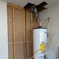 A white water heater stands in a wall alcove with exposed wooden studs, copper plumbing, and a vent pipe through the ceiling.