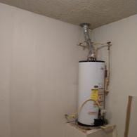 A white water heater stands in a corner with pipes connecting to the ceiling and wall in an unfinished room.