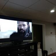 A TV screen displays a close-up of a bearded man with a serious expression, set in a dimly lit, furnished room.