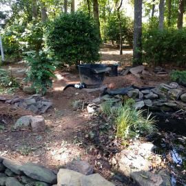 A shaded backyard landscape with a black garden pond filter, loose rocks, scattered plants, and trees in the background.