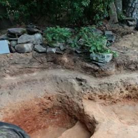 A muddy landscape showing an excavated trench with layered soil, rocky edges, and small green bushes in the background.
