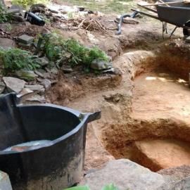 An excavation site shows deep pits dug into the earth, with a black plastic bin in the foreground and a wheelbarrow nearby.