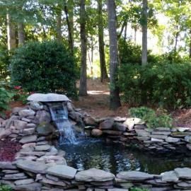 A stone-lined garden pond featuring a small waterfall, surrounded by lush green foliage and trees.