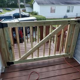 A wooden gate installed on a deck, viewed from behind, with a house and parked vehicles in the background.