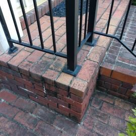Close-up of a brick porch step with a black metal railing base and a matching brick walkway below.