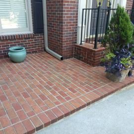A brick patio leads to a front door with a black iron railing, a turquoise ceramic pot, and a potted evergreen plant.