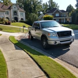 A silver Ford pickup truck parked on a suburban street next to a sidewalk with a hose extended to a nearby drainage grate.