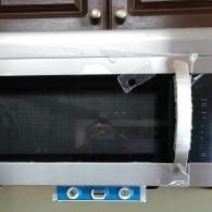 A silver microwave mounted under dark cabinets with a blue spirit level attached to the bottom.