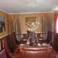 A dining room features a dark wood table with high-backed leather chairs, a floral centerpiece, and natural wall accents.