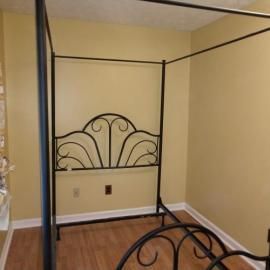 A black metal canopy bed frame with decorative arched headboard and footboard in a room with tan walls and wood floors.