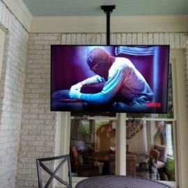 A flat-screen TV hangs from the ceiling in front of a brick wall and window, displaying a person in a mask and gloves.