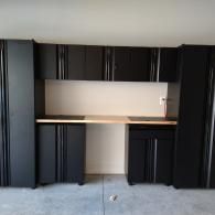 Black metal garage storage cabinets with a wooden countertop mounted on a wall against a plain light-colored background.