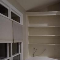 A window with light-colored blinds sits next to a built-in wall unit featuring several empty shelves.