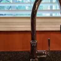 A chrome high-arc kitchen faucet stands on a dark granite countertop in front of a window with closed white blinds.