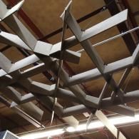 A sculptural, light-gray geometric baffle system hangs from a warehouse ceiling, creating a complex, interlocking pattern.