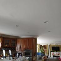 A view of a home kitchen with wooden cabinets and recessed lighting, looking into an adjacent living room.