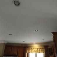 A low-angle view of a kitchen ceiling featuring recessed lighting above wooden cabinets and a window with a valance.