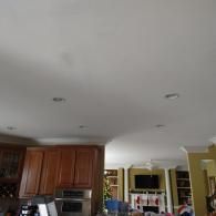 A kitchen with wood cabinetry and recessed lighting, looking toward a living area with a television above a fireplace.