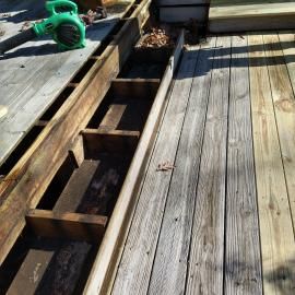 A partially dismantled wooden deck showing exposed joists and framing, with a leaf blower nearby on the left.