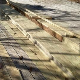 A wooden deck with weathered planks in various states of gray and brown, crossed by diagonal shadows.