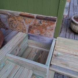 A wooden deck hatch is open, revealing a square storage compartment against a stone-accented green exterior wall.