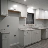 A modern kitchen with white shaker-style cabinets, grey countertops, and light grey walls under recessed lighting.
