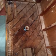A high-angle view of a bathroom subfloor under renovation, featuring diagonal wooden planks and a central toilet drain pipe.