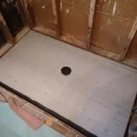 A concrete shower floor base with a central drain, framed by wooden wall studs during construction.