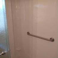 A brushed metal grab bar is installed horizontally on a plain white shower wall, with a glass shower door on the left.